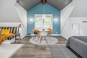 A bedroom with a sloped ceiling, a bed, a window with blinds, a dining table with chairs, and a sofa with cushions. - Carriage House - Studio Condo minutes away from downtown Saugatuck! (Saugatuck)
