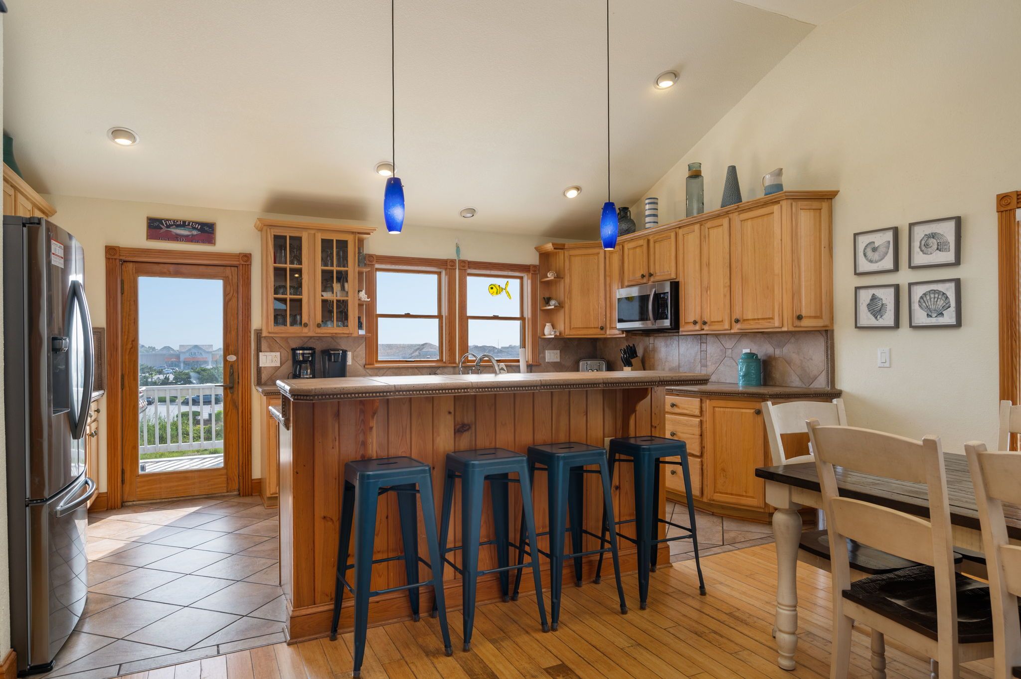 A kitchen with wooden cabinets, a central island with stools, a refrigerator, and a view through the windows.