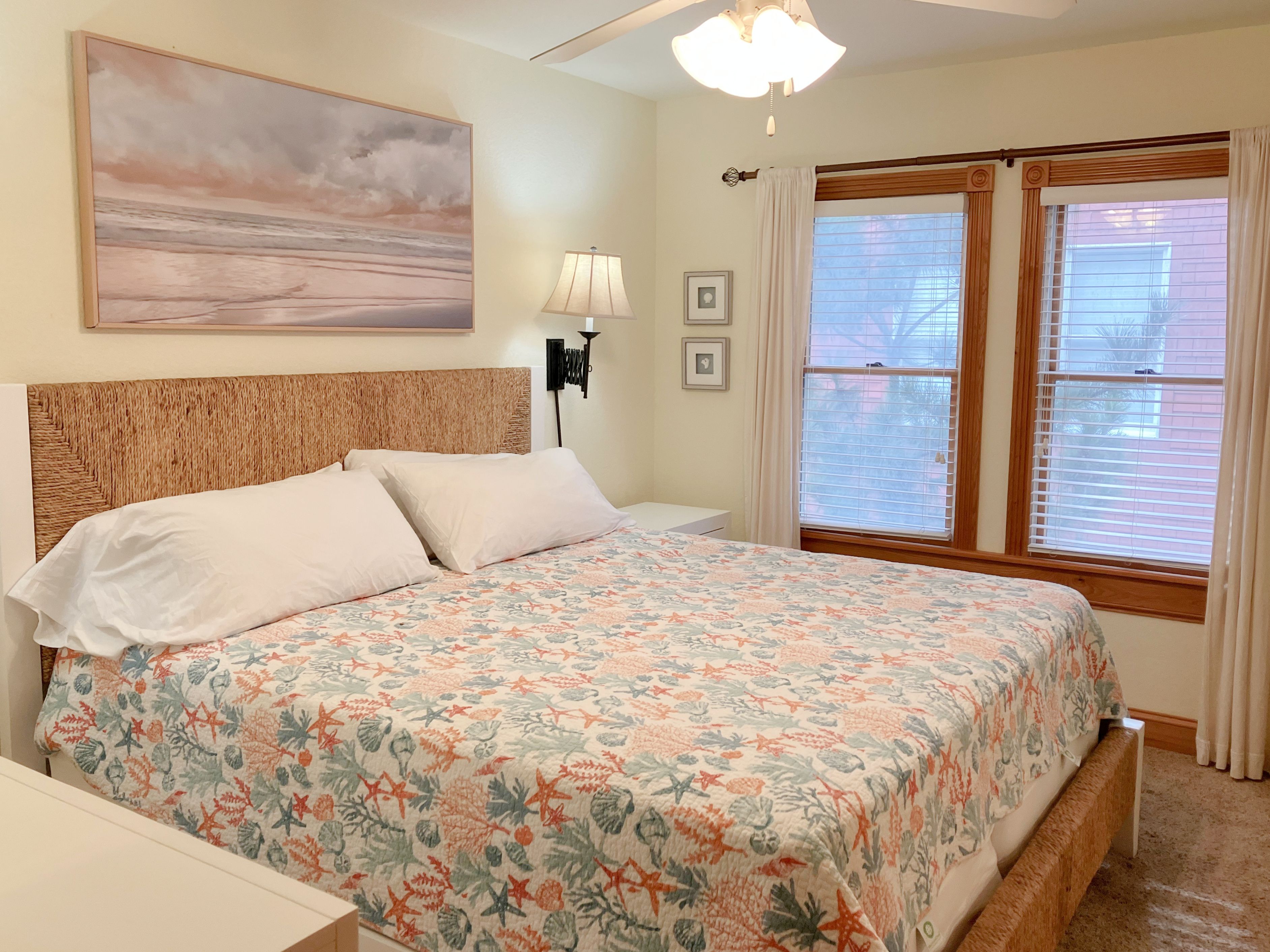 A bedroom with a bed, a nightstand, a lamp, a window with blinds, and a wall-mounted picture.