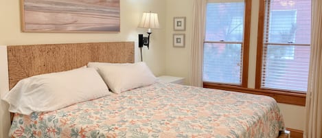 A bedroom with a bed, a nightstand, a lamp, a window with blinds, and a wall-mounted picture.