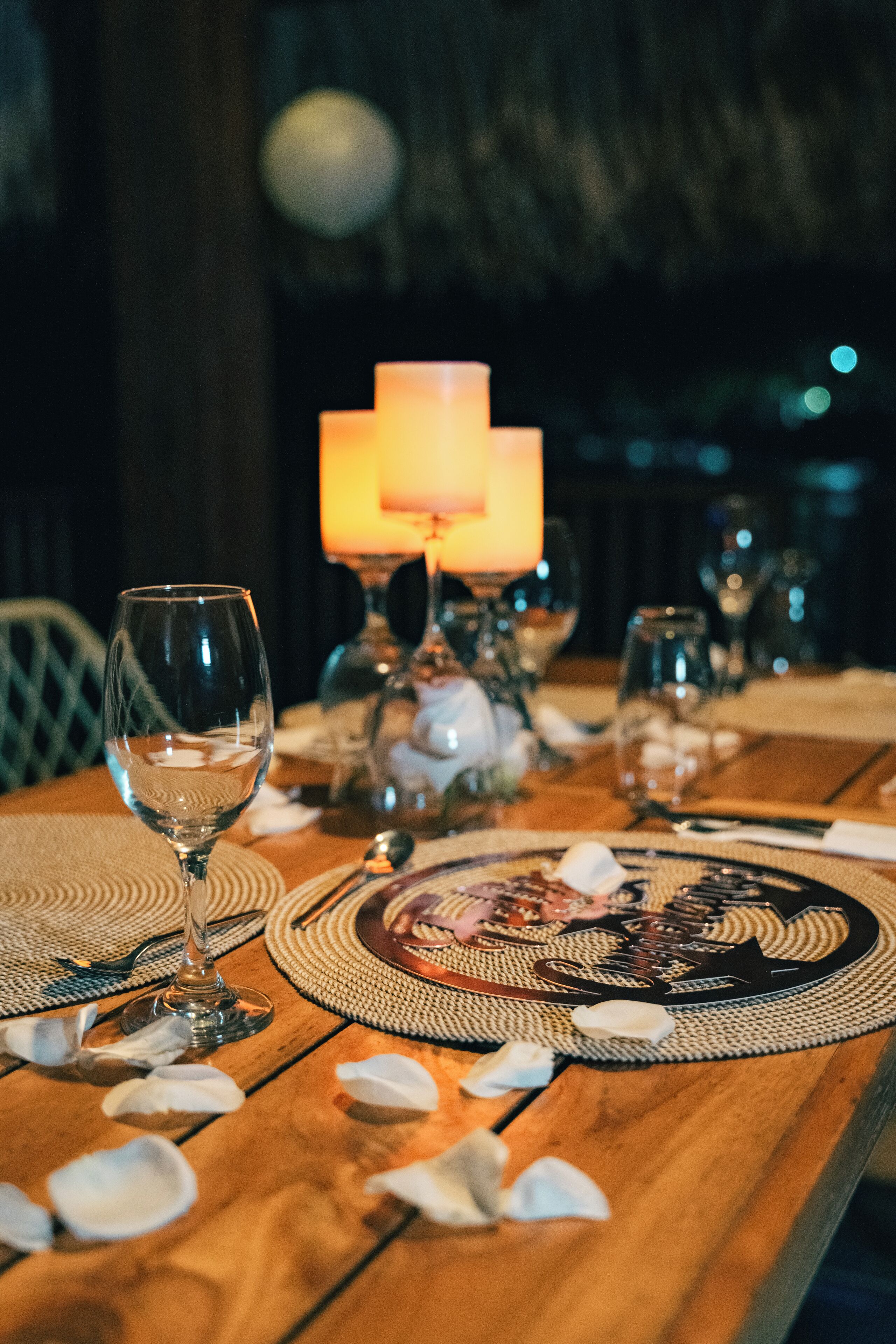 Mesa de madeira posta com taças de vinho, velas e pétalas de rosas espalhadas.