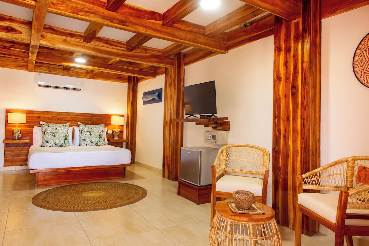 A bedroom with a wooden ceiling, a bed with floral pillows, a TV, a refrigerator, and wicker chairs.