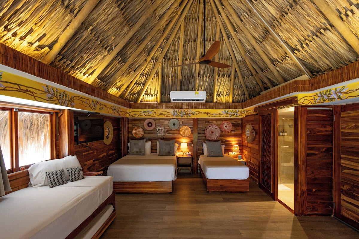 A room with two beds, wooden walls, a thatched ceiling, and a central air conditioning unit.