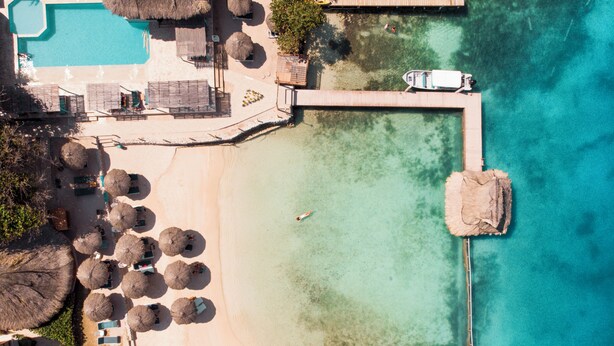 An aerial view of a beach resort with a swimming pool, umbrellas, lounge chairs, and a pier extending into the water. - Hotel Isla del Sol  (Rosario Islands)