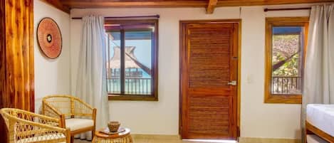 Una habitación con vigas de madera, una puerta grande de madera, dos ventanas con cortinas, muebles de mimbre y una cama.