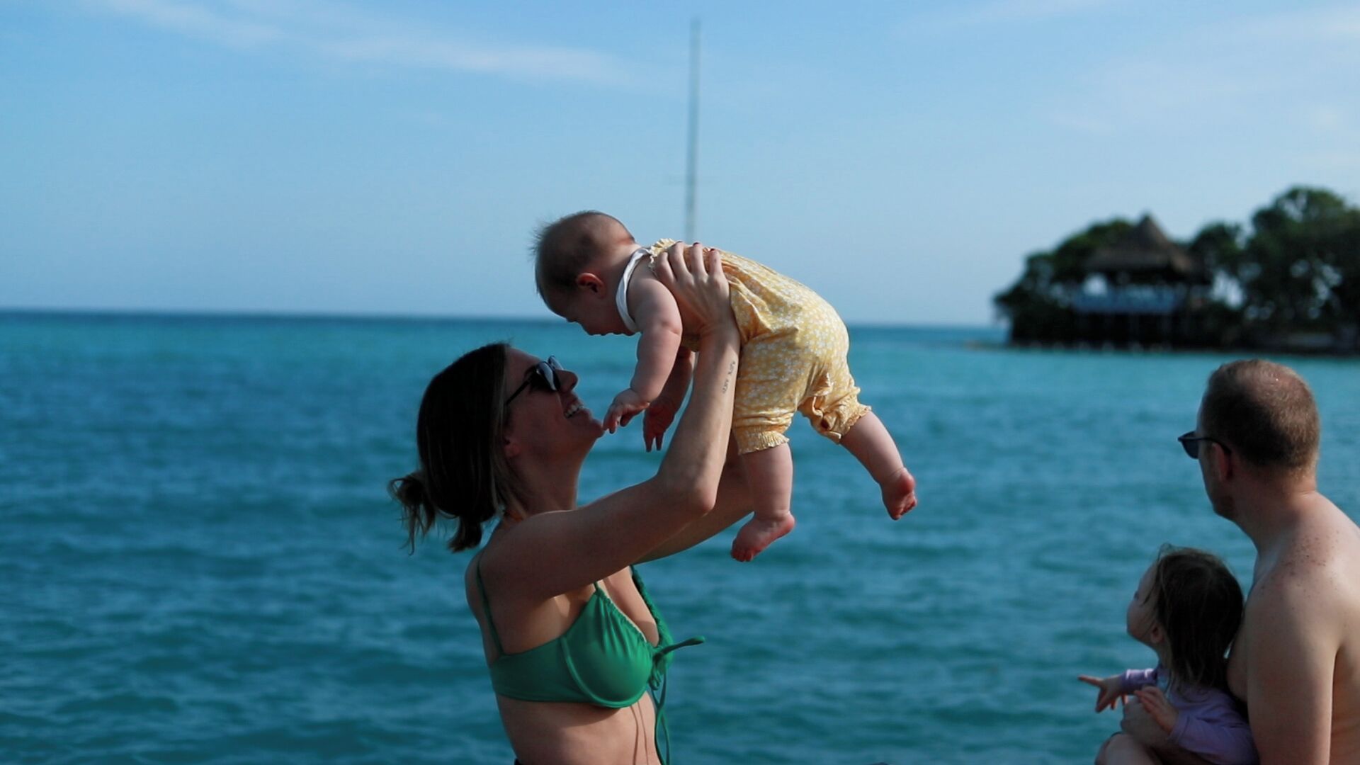 Una persona sosteniendo a un niño sobre una masa de agua, con una estructura distante en el horizonte.
