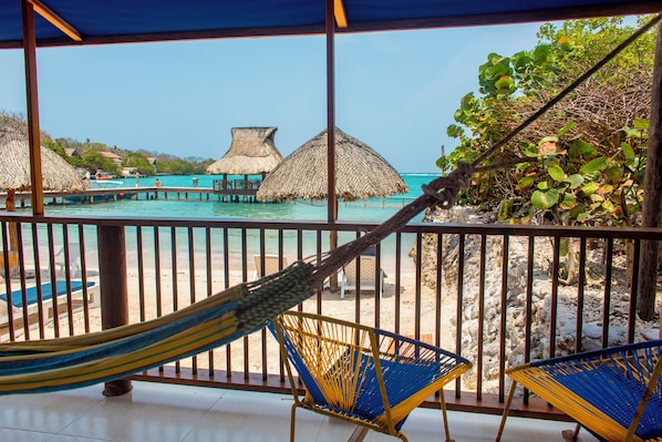 A beach view from a covered area with a hammock, chairs, and thatched umbrellas. - Hotel Isla del Sol (Rosario Islands)