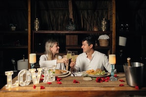 A couple toasting with drinks at a table with 'LOVE' spelled out in lights, surrounded by rose petals and candles, in a dimly lit room with wooden shelves and a rustic ambiance.