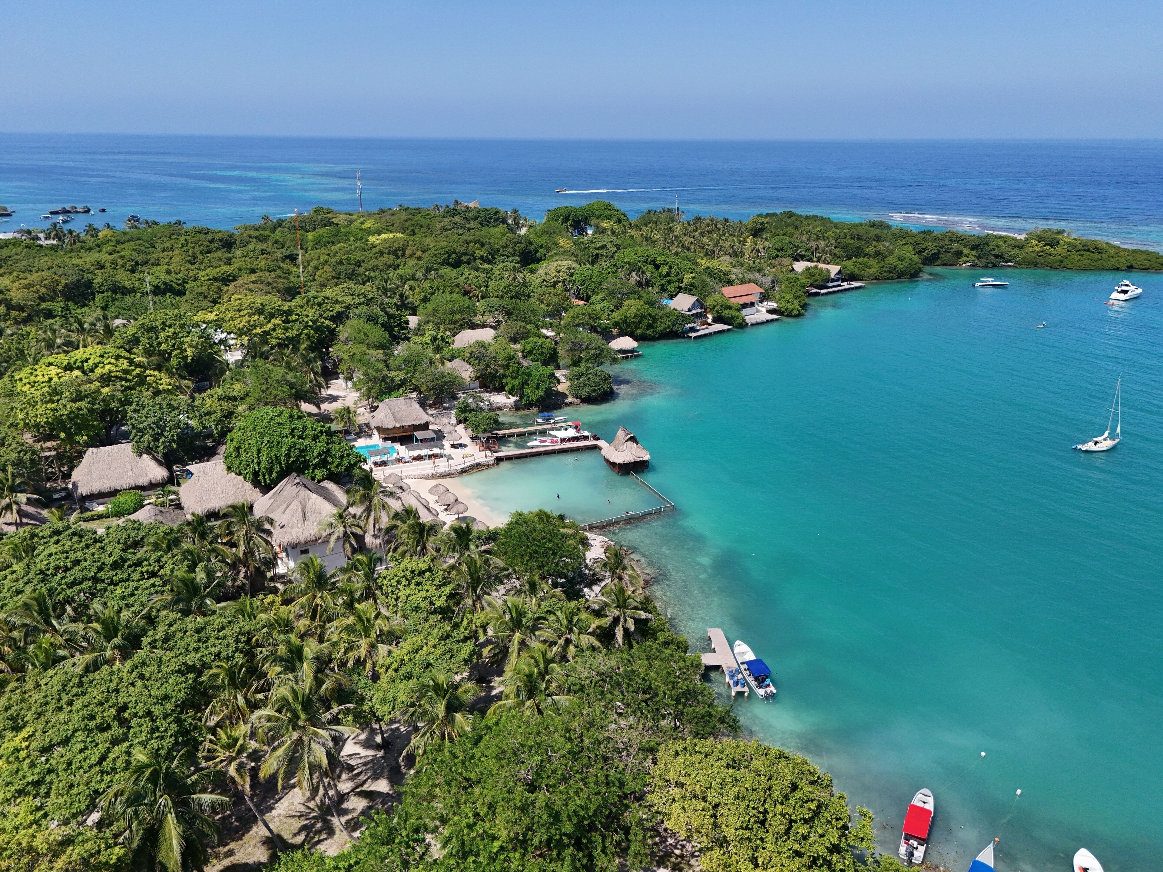 An aerial view of a tropical island with clear turquoise waters, a dock with boats, and thatched-roof huts.
