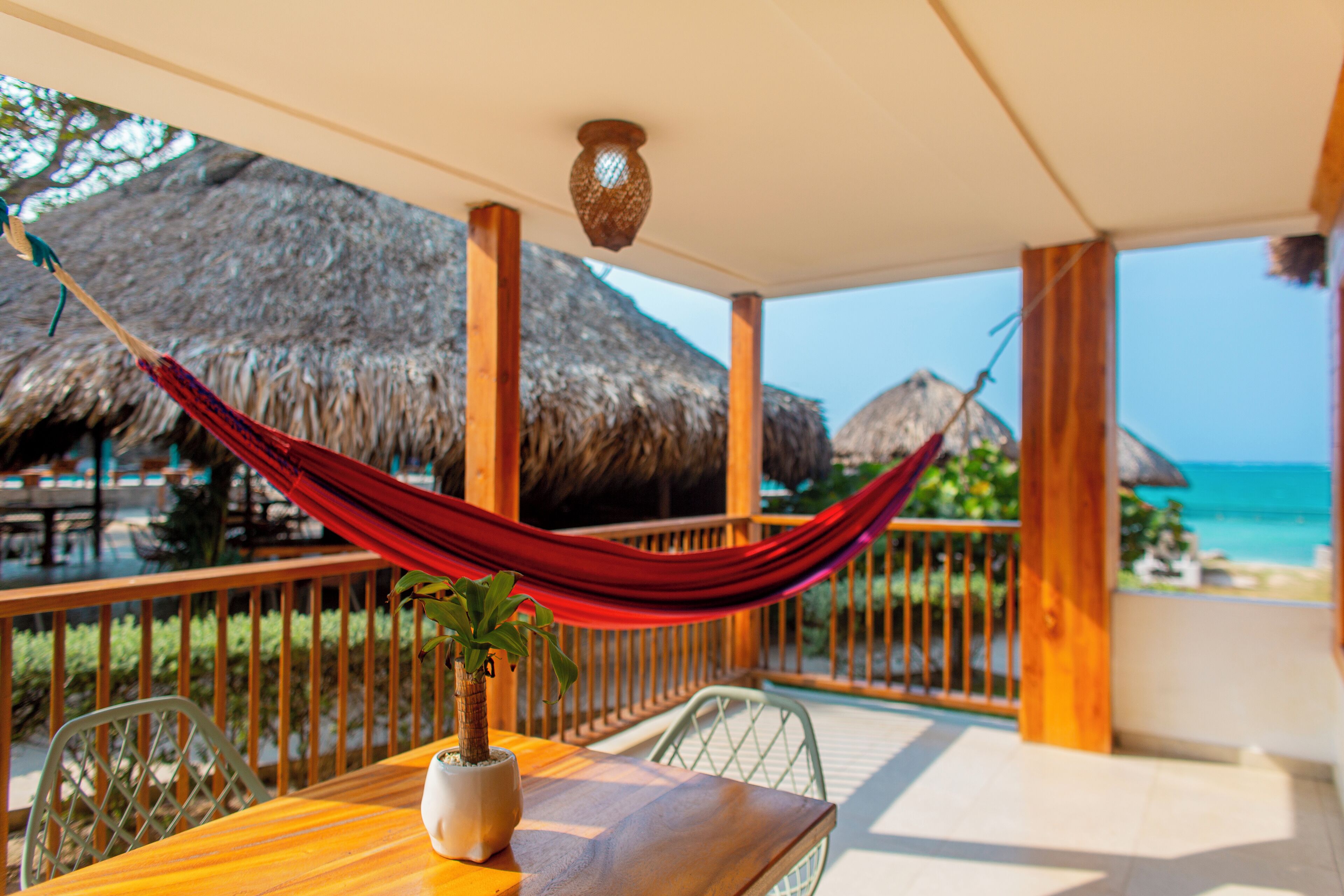 A wooden table with a potted plant, a hammock, and a view of the ocean and thatched-roof structures.