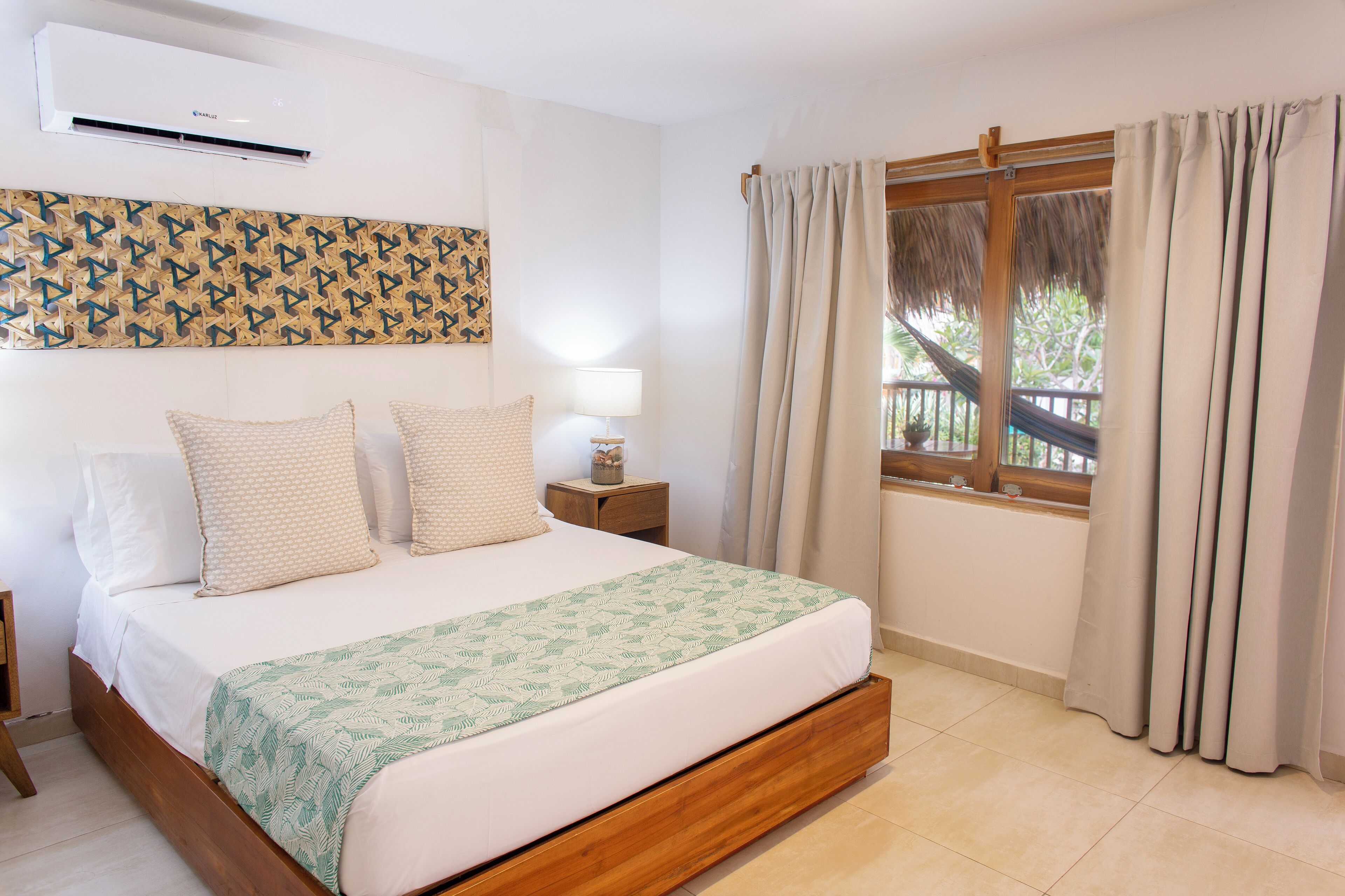 A bedroom with a bed, a nightstand, a lamp, a window with curtains, and a wall-mounted air conditioner.