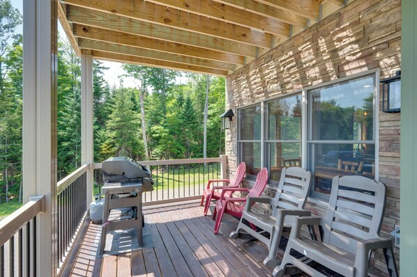 A covered deck with Adirondack chairs, a grill, and a view of a wooded area.