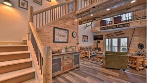 Ein rustikales Blockhaus-Interieur mit einer Holztreppe, einem Essbereich mit Tisch und Stühlen, einer Couch und einem Schild mit der Aufschrift 'Back Up Ta Camp'.