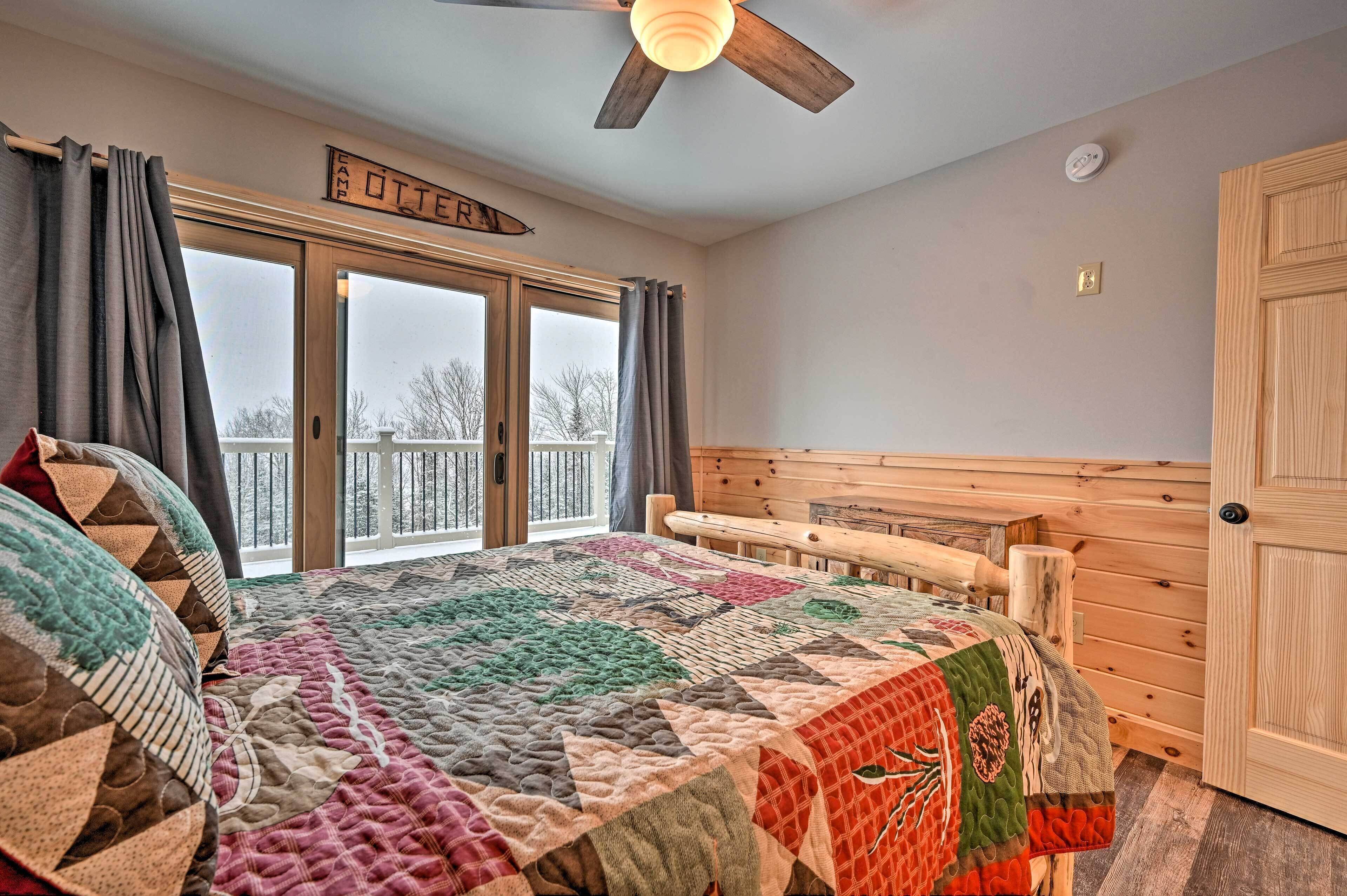 A cozy bedroom with a large bed, a wooden headboard, a ceiling fan, and a view of a snowy landscape through a sliding glass door.