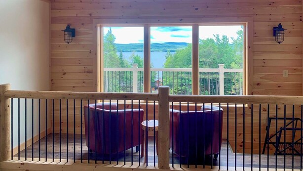 Eine Holzhütte mit Balkon und Blick auf einen See, möbliert mit roten Stühlen und einem kleinen Tisch.