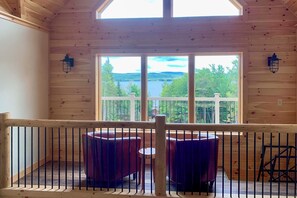A wooden cabin with a balcony overlooking a lake, furnished with red chairs and a small table.