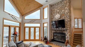 A spacious living room with a stone fireplace, large windows, and a high ceiling.