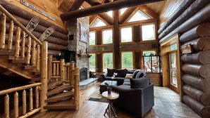 A log cabin interior with a stone fireplace, a black leather sofa, and a wooden staircase. - WM dream home in National Forest; AC, swimspa, beach, massage chair, 2 min from ski area! (Bretton Woods)