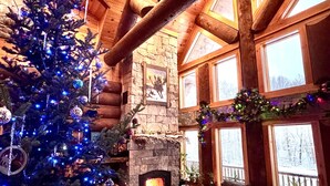A cozy log cabin interior with a decorated Christmas tree, a stone fireplace, and a large window overlooking a snowy landscape. - WM dream home in National Forest; AC, swimspa, beach, massage chair, 2 min from ski area! (Bretton Woods)