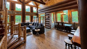 A log cabin interior with a large wooden staircase, a sectional sofa, a dining area, and large windows offering a view of the forest. - WM dream home in National Forest; AC, swimspa, beach, massage chair, 2 min from ski area! (Bretton Woods)