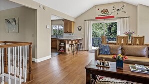 Un salon spacieux doté d’un plancher en bois, d’un canapé, d’une table basse avec un jeu d’échecs, et offrant une vue sur le jardin arrière grâce à de grandes fenêtres.