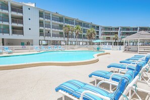 A swimming pool with lounge chairs, a multi-story building with balconies, and a beach area.