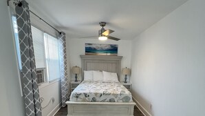 A bedroom with a bed, a ceiling fan, a window with blinds, and a painting of a beach scene. - Crystal Sands Apartments  (Ocean City)