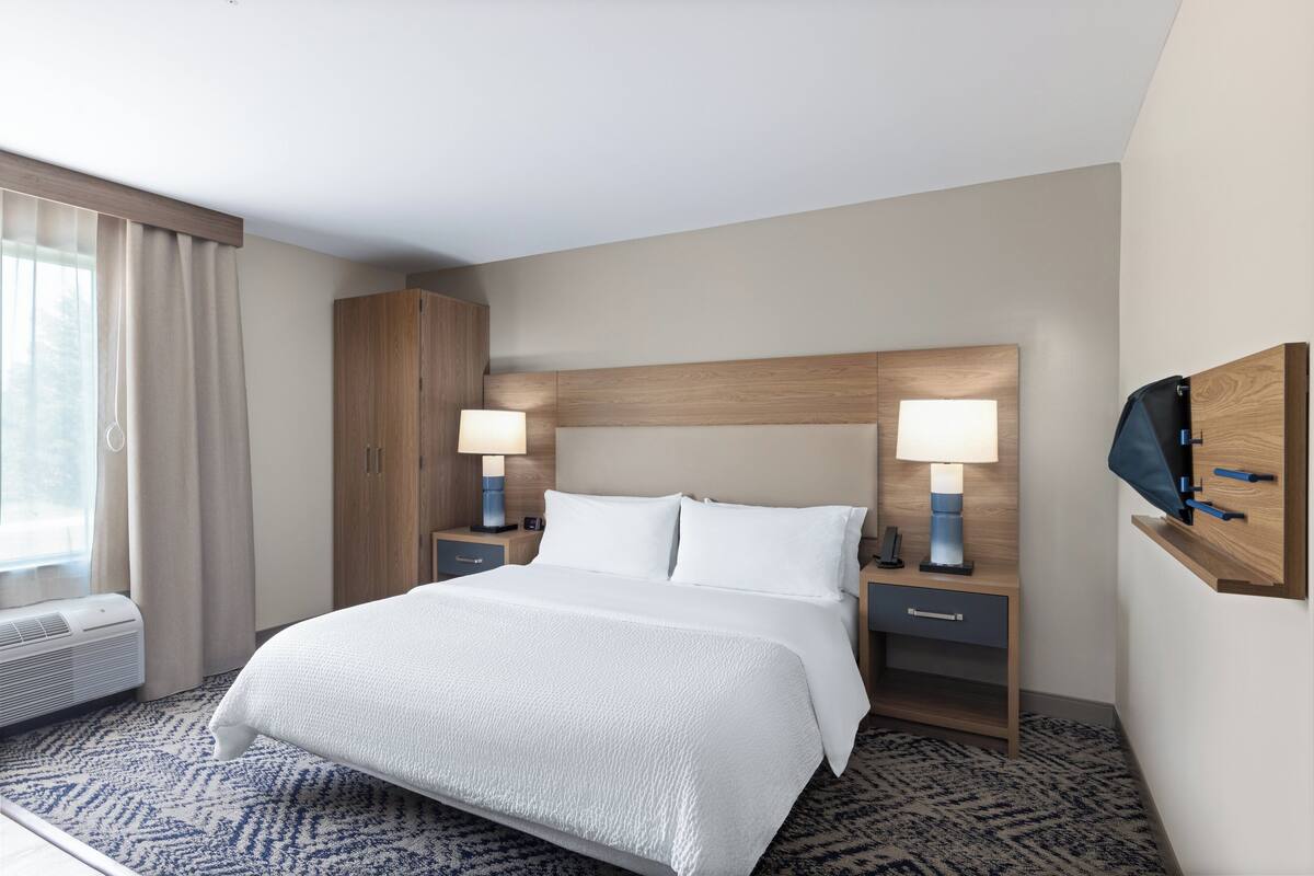 A hotel room with a large bed, two bedside tables with lamps, a wardrobe, an air conditioning unit, and a TV mounted on the wall.
