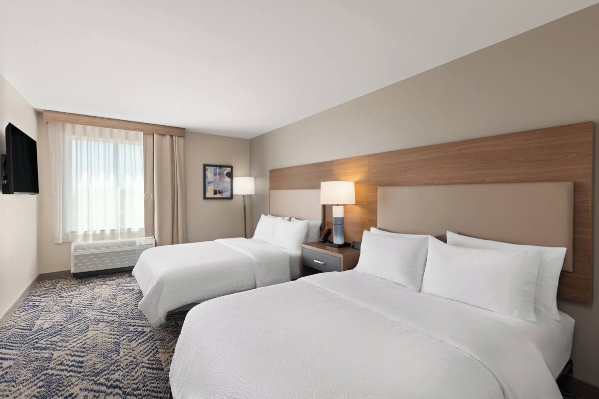 A hotel room with two beds, a flat-screen TV, a bedside table with a lamp, and a large window with curtains.