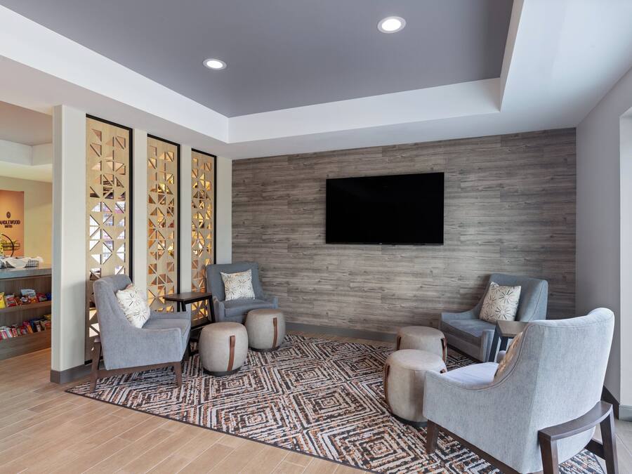 A modern living area with a flat-screen TV mounted on a wooden wall, grey upholstered chairs, a patterned rug, and a snack bar with a variety of items.