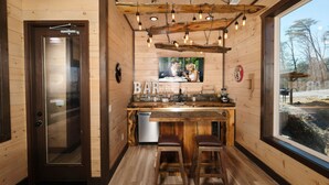 A cozy wooden cabin with a bar area featuring a rustic kitchen, wooden stools, and a large window offering a view of the outdoors. - Splashin' Mountain Retreat - Sip Cocktails Fireside, Swim with Fountains & Project Movies! (Sevierville)