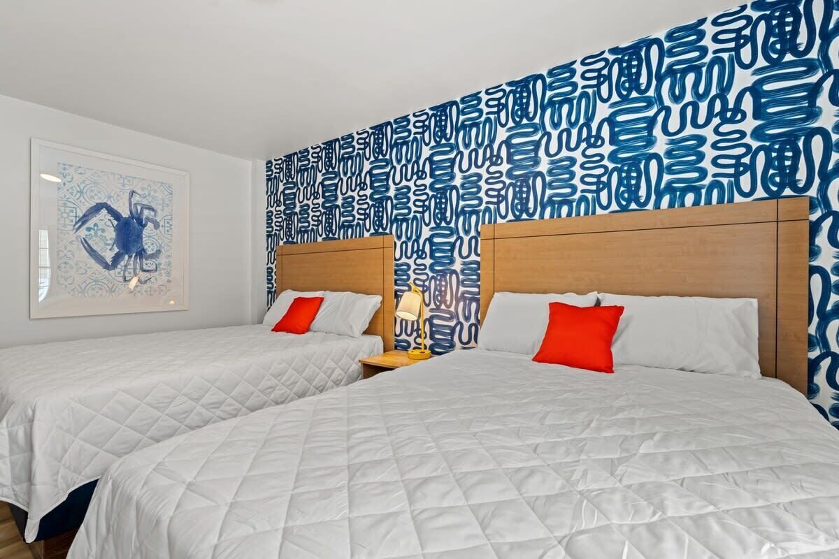 Ein Hotelzimmer mit zwei Betten, weißer Bettwäsche, roten Kissen, einem hölzernen Kopfteil, einer Wand mit blauem abstrakten Muster und einem Bilderrahmen an der linken Wand.