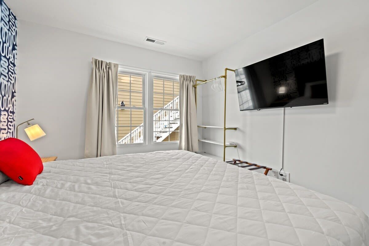 Ein Schlafzimmer mit einem großen Bett, einem an der Wand montierten Fernseher, einem Fenster mit Vorhängen und einer durch das Fenster sichtbaren Treppe.