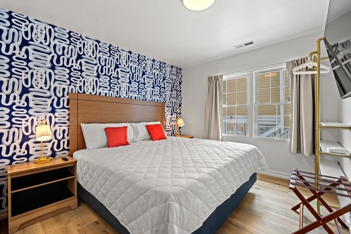 Une chambre d’hôtel avec un grand lit, une table de chevet avec une lampe, une fenêtre avec des rideaux et un mur décoré d’un motif.
