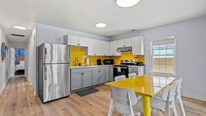A modern kitchen with white cabinets, a stainless steel refrigerator, a yellow dining table, and a window with blinds.