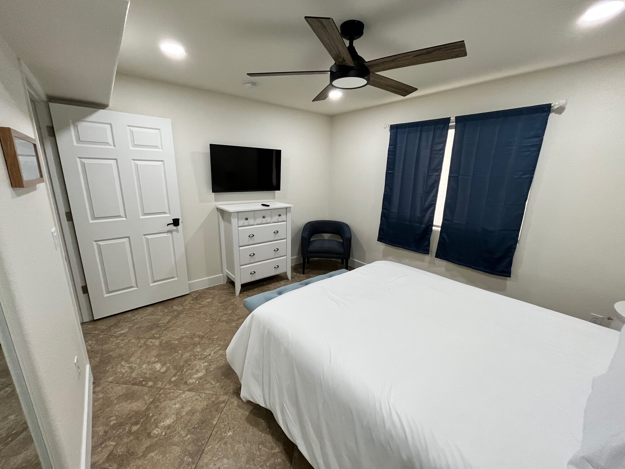 Un dormitorio con una cama, un aparador, una silla, un ventilador de techo y un televisor.