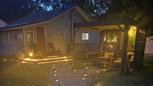 Una casa con veranda e area salotto esterna, un vialetto che conduce all'ingresso e una piccola terrazza con luci decorative.