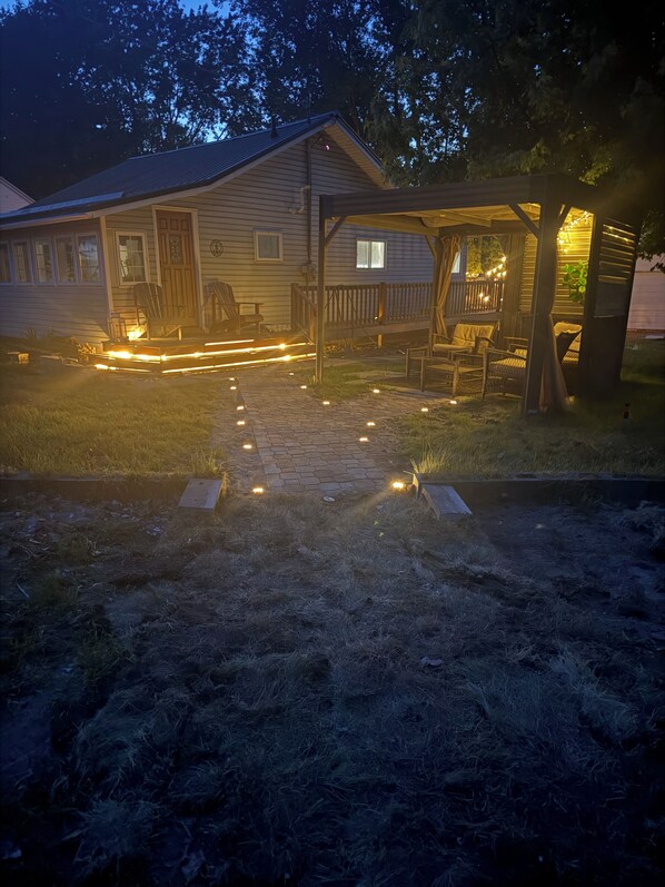 A house with a porch and outdoor seating area, a pathway leading to the entrance, and a small deck with string lights. - Newly updated cottage with new central air just steps from the beach (Port Rowan)