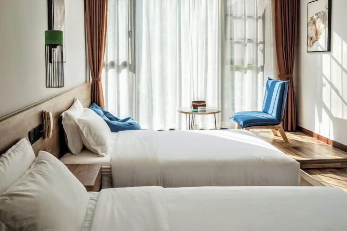 A hotel room with two beds, a small round table, a blue chair, and a window with sheer curtains.