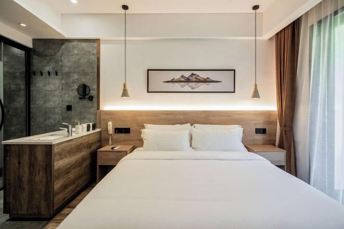 A modern hotel room with a wooden headboard, a large bed, a built-in vanity with a mirror, and a framed artwork above the bed.