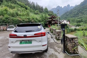 A white car parked at a charging station in a rural setting with a traditional building in the background. - Mini Inn (Zhangjiajie)