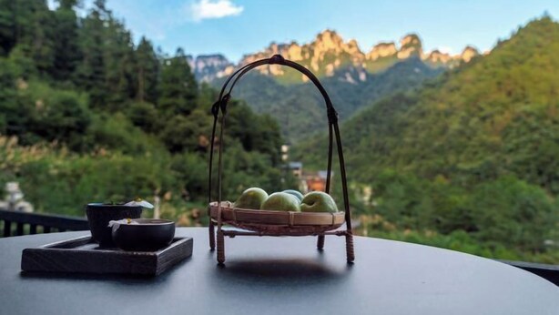 A table with a basket containing apples, a bowl, and a cup, set against a mountainous backdrop. - Mini Inn (Zhangjiajie)