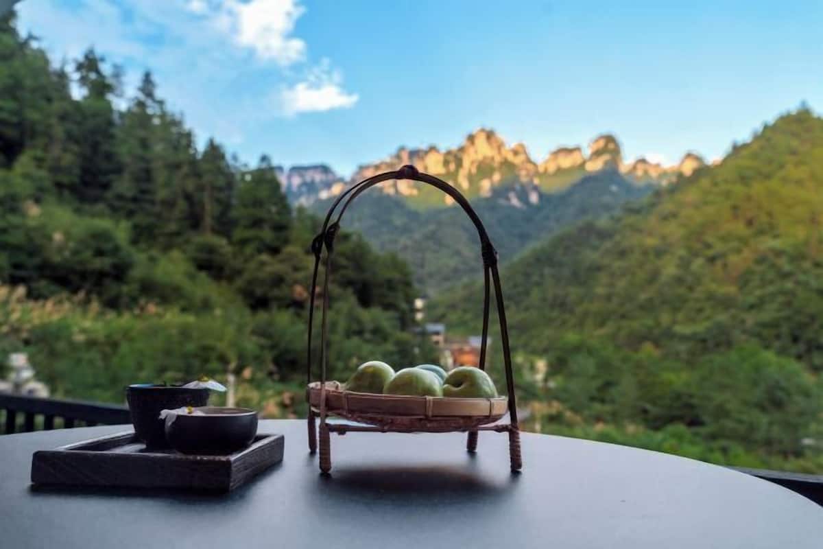 A table with a basket containing apples, a bowl, and a cup, set against a mountainous backdrop.