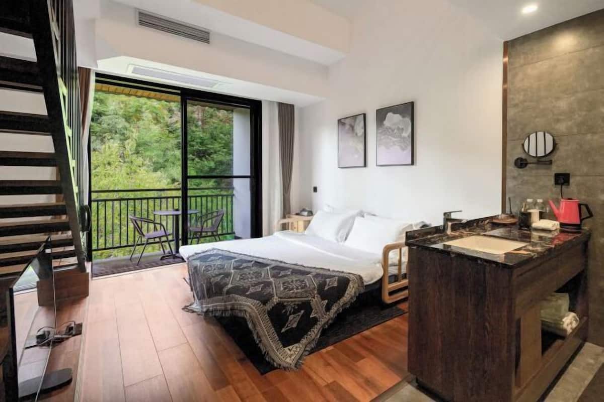 A bedroom with a bed, a wooden floor, a balcony with a chair, and a small kitchen area.