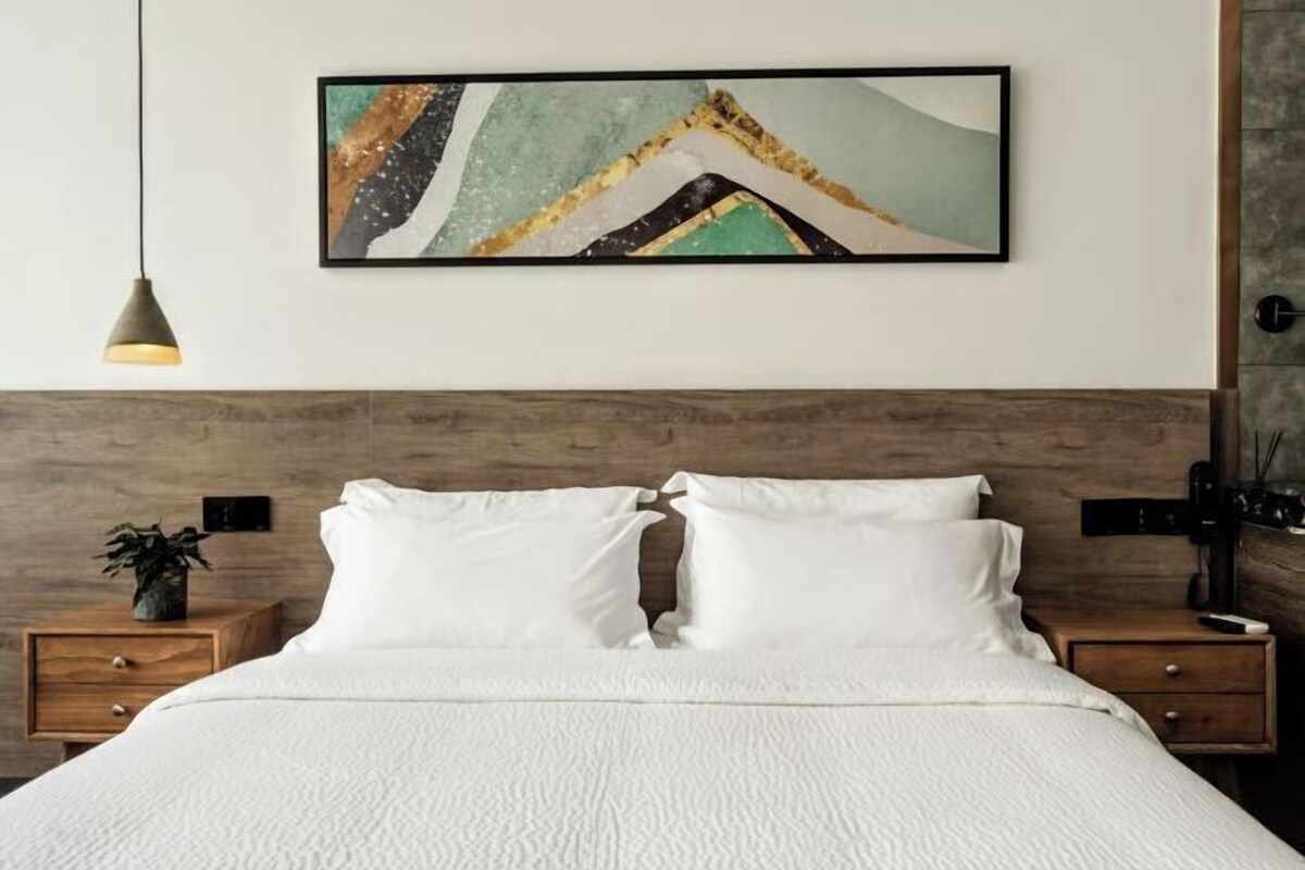 A neatly made bed with white bedding, a wooden headboard, and a wall-mounted abstract painting.