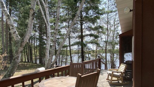 Une terrasse en bois avec une table et des chaises, donnant sur un lac et entourée d’arbres.