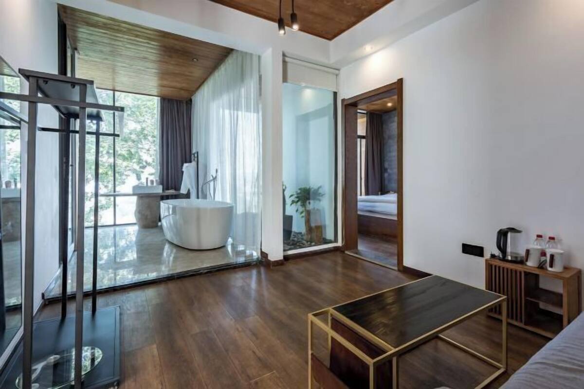 Una habitación de hotel moderna con suelo de madera, cabina de ducha de vidrio, bañera blanca y una mesita de noche de madera.