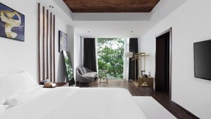 Una habitación de hotel moderna con una cama grande, un área de descanso con una silla y mesa, un televisor y un balcón con vistas a árboles.