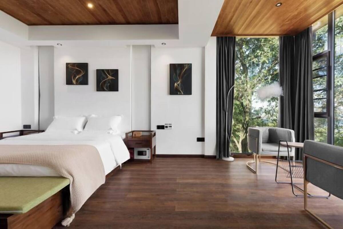 Habitación de hotel moderna con techo de madera, cama con un banco verde, escritorio y silla. Hay pinturas abstractas en la pared y vistas a árboles a través de amplias ventanas.