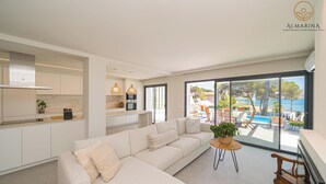 En moderne stue med en beige sofa, et lille rundt bord og udsigt til pool og strand gennem store vinduer.